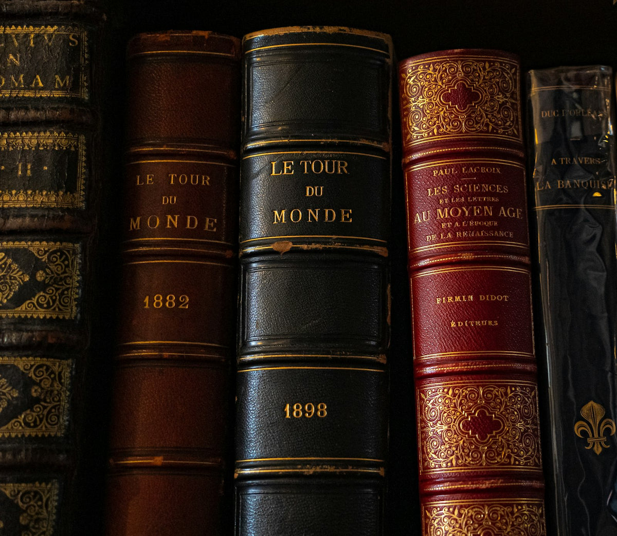A row of worn antique books with aged, sun-faded spines on a wooden library shelf, lit by soft natural daylight
