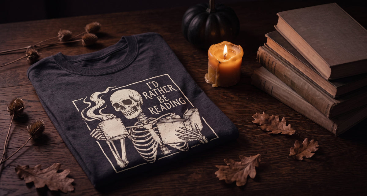 An I'd Rather Be Reading tee folded on a dark table beside a lit candle and a stack of old books